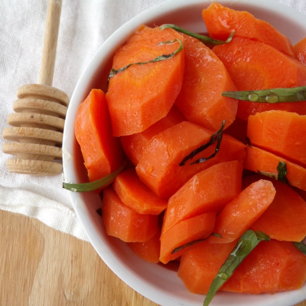 A white bowl filled with carrots, topped with tarragon and honey sitting on top of a white towel with a honey drizzler.