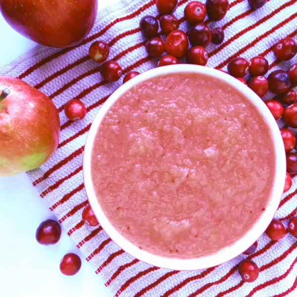 A bowl of Cranberry applesauce sitting on a white and red striped towel with apples and cranberries next to it.
