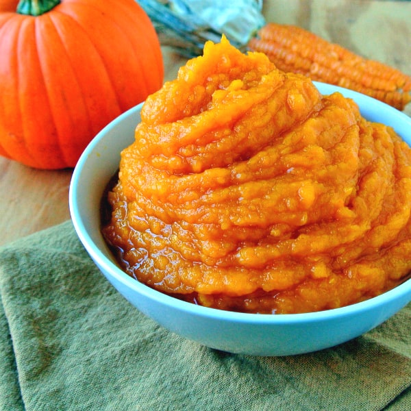 A bowl of Alton Brown Pumpkin Puree with a pumpkin and a corn cob behind it.