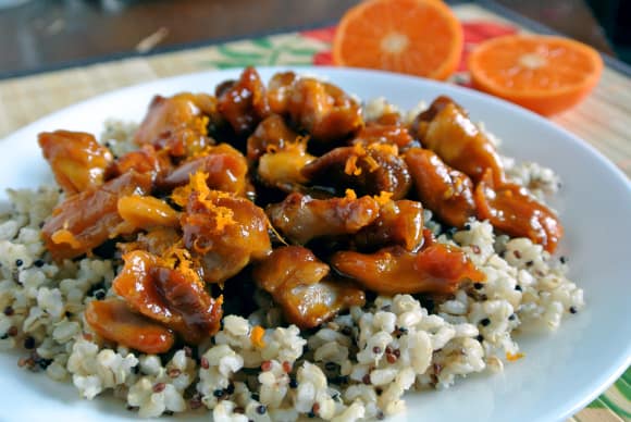 Clementine glazed chicken on top of a rice-quinoa medley on a white plate with a cut in half clementine in the background. 