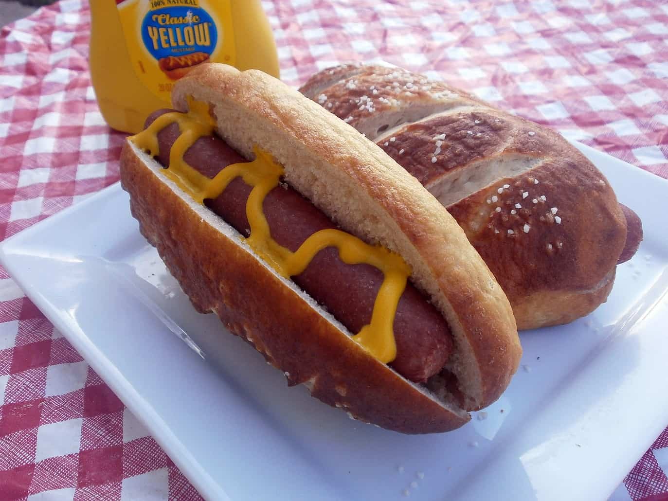 Two soft pretzel buns with hot dogs inside topped with mustard sitting on a white plate.