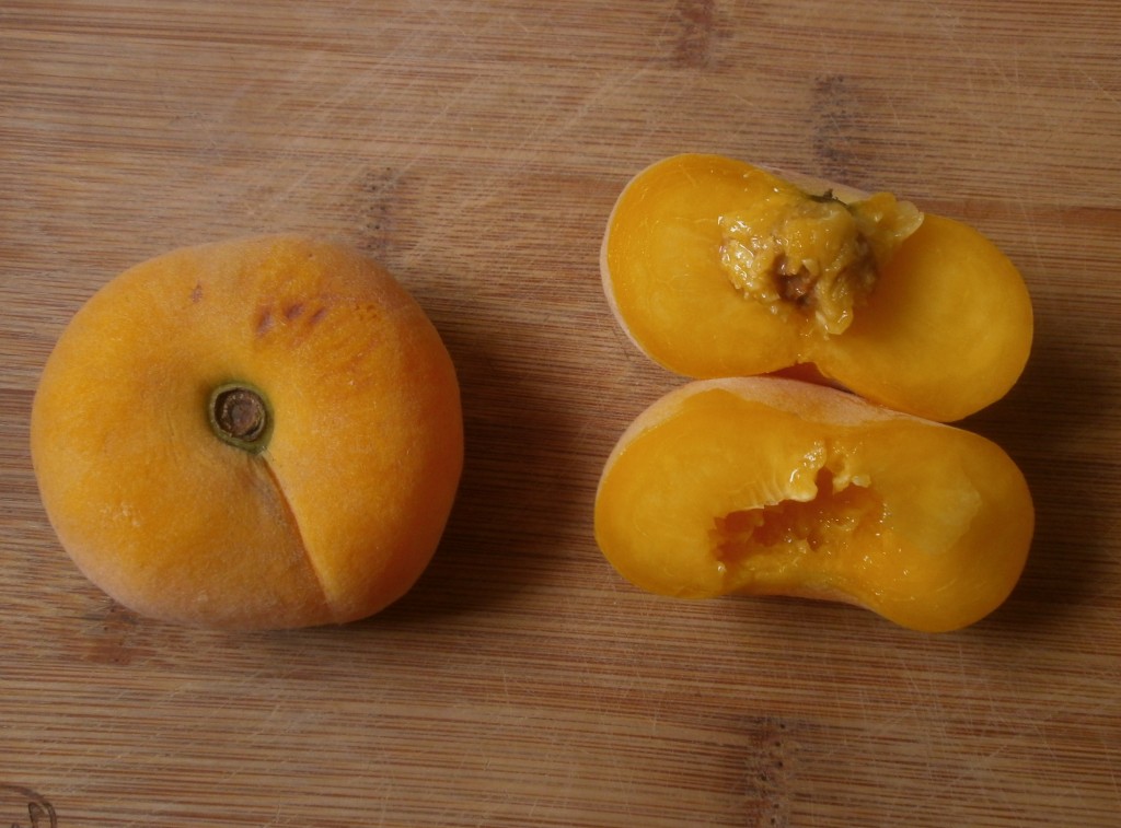 A whole yellow Peach Pie donut peach next to one that has been cut in half to show the small pit inside and that the flesh is the same color as the skin. The peaches are sitting on a wood cutting board.