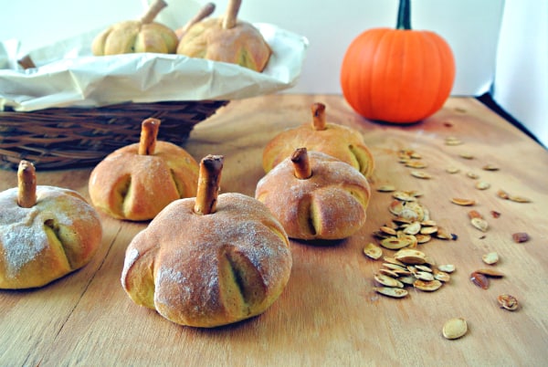 Pumpkin Yeast Rolls shaped into mini pumpkins with a pretzel on top for a steam are sitting on a wooden board with roasted pumpkin seeds and a whole pie pumpkin in the background.