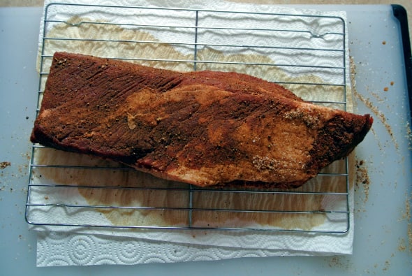 A seasoned brisket is shown on a cooling rack on top of a cutting board with a paper towel after having been in the fridge for 24 hours. The meat has darkened in color.