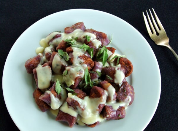 Browned purple sweet potato gnocchi covered in a white cheese sauce and fresh sage on top of a white plate on top of a black background with a fork next to the plate.
