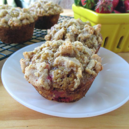 A white plate with 2 Strawberry Rhubarb Muffins with more of them in the background on a cooling rack and yellow container of fresh strawberries. 