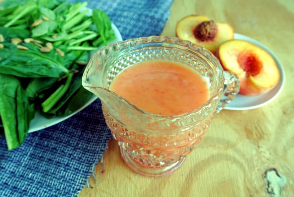 A glass jar with a peach vinaigrette inside sitting on a wood board with a salad on a plate next to it and a sliced peaches on a plate behind it.