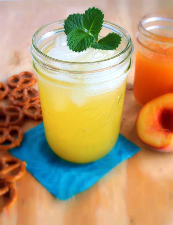 A ball jar with Homemade Peach Soda Pop in it surrounded by peaches and pretzels.