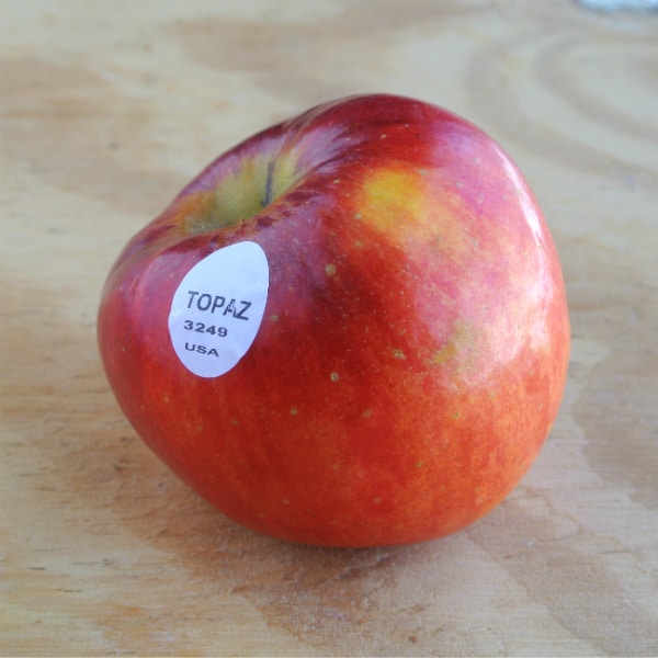 A single Topaz apple sitting on a wood board. The plu sticker on shown on the fruit saying "Topaz 3249 USA".