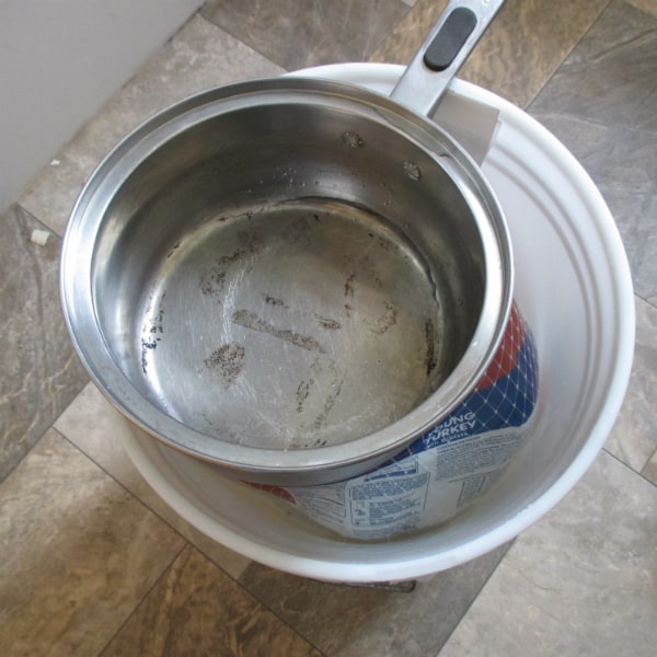 A frozen turkey in it's packaging inside a 5 gallon white bucket filled with water sitting on the floor. A pot is on top of it to keep the turkey covered in water.