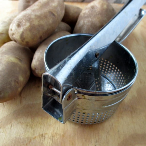 A metal potato ricer on a wood board in front of some russet potatoes