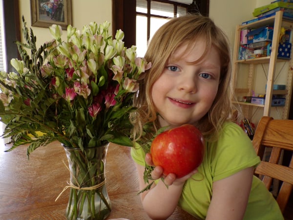 My daughter sitting at a table holding a Sugar Bee apple next to a vase of cut flowers.