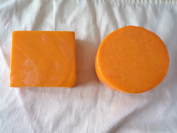 A square block of cheddar cheese next to a circle of colby cheese on a white towel.