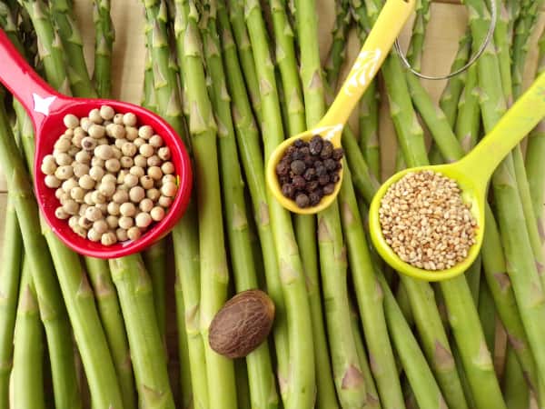 3 spoons one with sesame seeds, one with white peppercorns, one with black peppercorns, and nutmeg by itself on top of asparagus