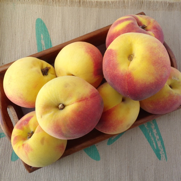 A wood tray full of peaches sitting on top of cloth placemat that is brown with streaks of turquoise. 