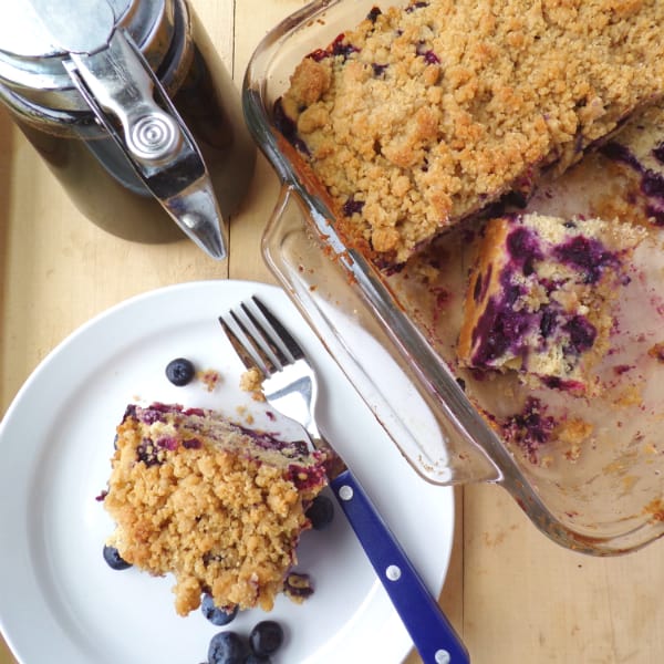 Blueberry Buckle slice on a plate with the buckle in a glass pan next to it. A bottle of maple syrup is located nearby as well.