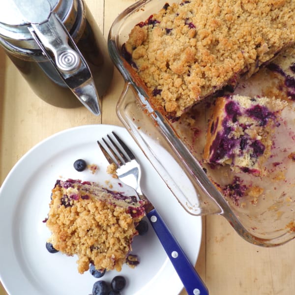 Blueberry Buckle slice on a plate with the buckle in a glass pan next to it. A bottle of maple syrup is located nearby as well.