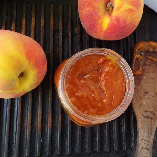 A mason jar full of peach bbq sauce on top of a cast iron grill pan. Peaches are on top of the grill pan and a wood spatula is next to the jar.
