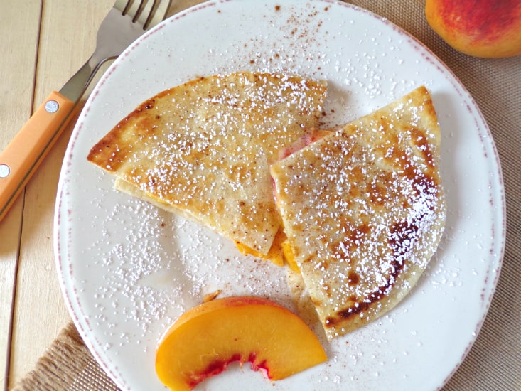 Slices of peach quesadilla sitting on a white plate with brown trim. The quesadilla are filled with peaches and Mascarpone cheese and sprinkled with powdered sugar.