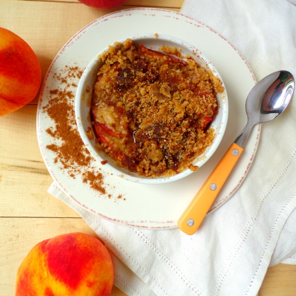 A single serving peach cobbler in a white ramekin on a white plate with brown trim. A spoon with a orange handle is on the plate and a whole peach is to the left and underneath.