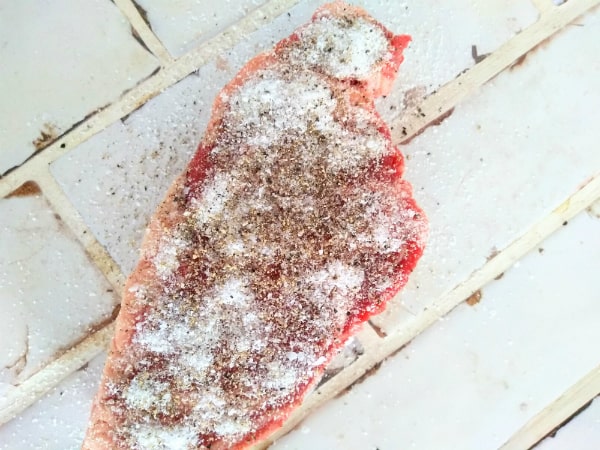 A raw center eye chuck eye steak sitting on white tile countertop seasoned with kosher salt and freshly cracked black peppercorns.