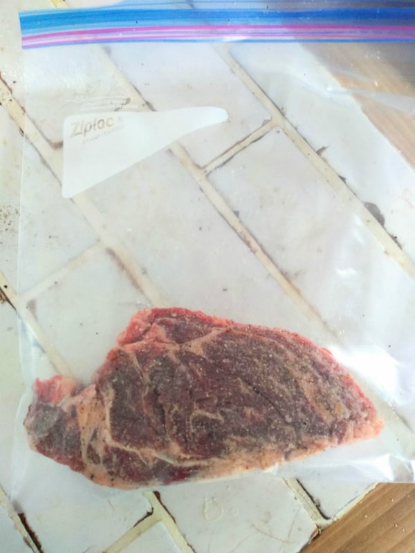 A Ziploc freezer bag with a chuck eye steak inside of it on a white tile countertop.