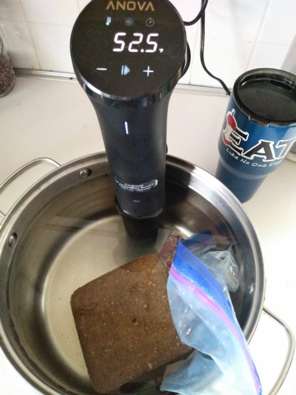 A stock pot with an Anova sous vide machine attached to it. The pot is filled with water and a brick is being used to keep the steak in a gallon zip top bag under water.