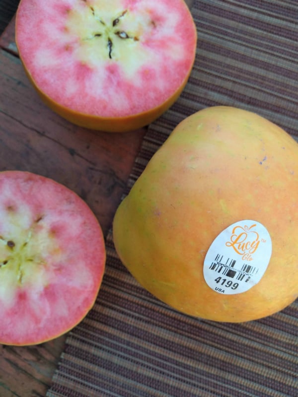 A Yellow Skinned apple with a Lucy Glo sticker on a brown placemat on a brownish red table. One apple is split in half showing the pink inferior.