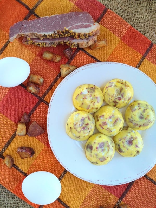 A white plate with egg bites containing leftover corned beef and potatoes, sitting on a orange plaid towel with whole egga next to the plate.