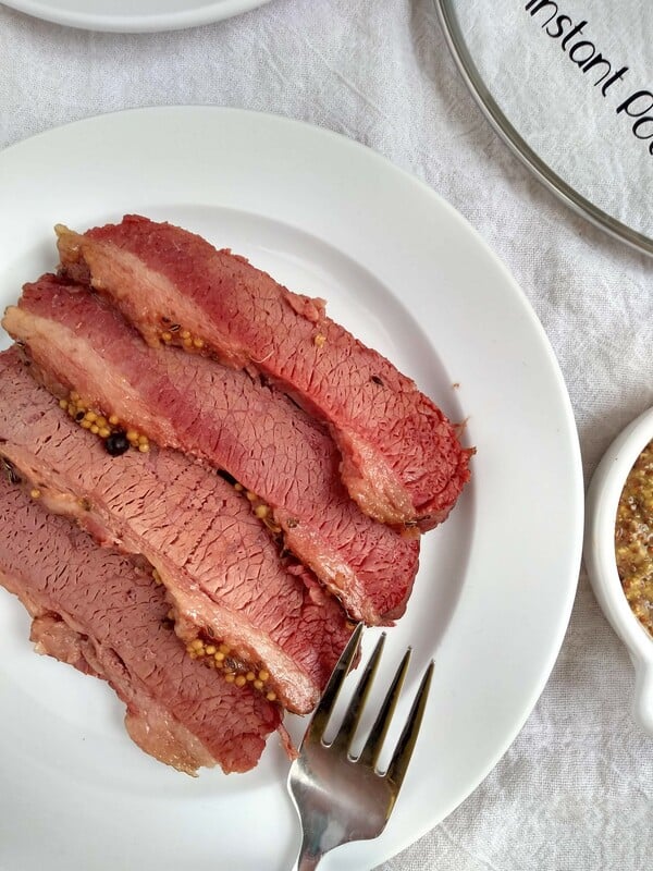 Sliced pieces of corned beef on a white plate on white towel with a glass Instant Pot lid next to it.