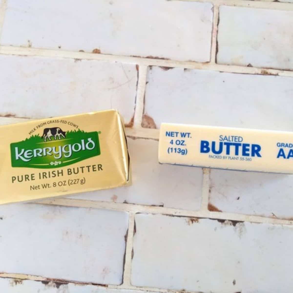 A 8 ounces package of Kerrygold Pure Irish Butter next to a store brand salted butter 4 oz stick. They are sitting on top of a white tile counter top.