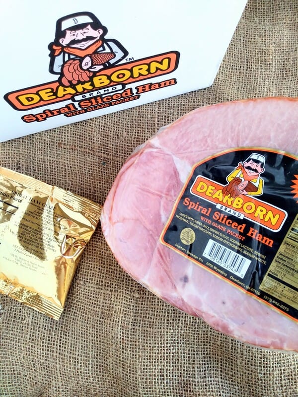 A Dearborn Spiral Sliced Ham sitting on on burlap with the sauce packet next to it and a white Dearborn brand box.