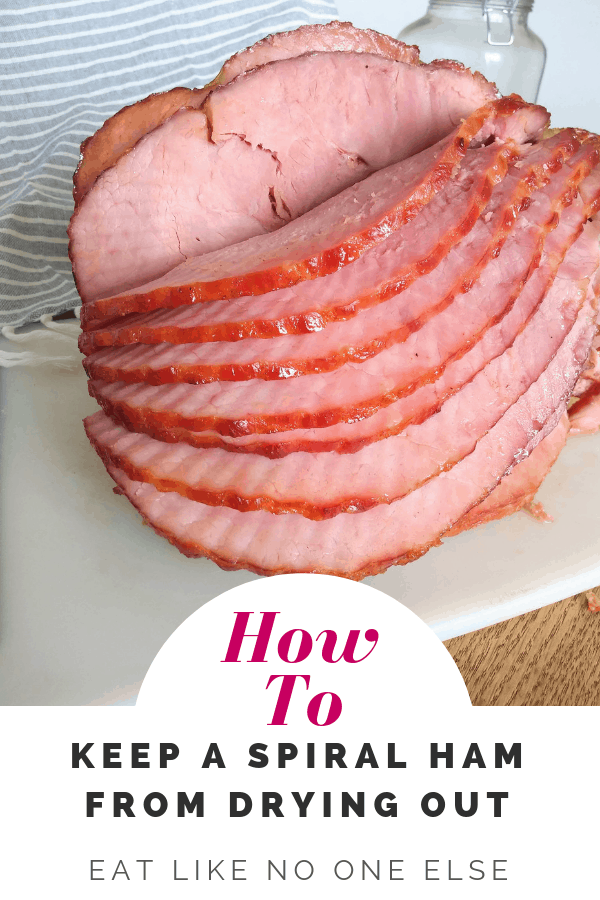 A spiral sliced ham on a white cutting board on a wood table with a gray striped towel in the background. The words "How to Keep a Spiral Ham From Drying Out" are below the picture.