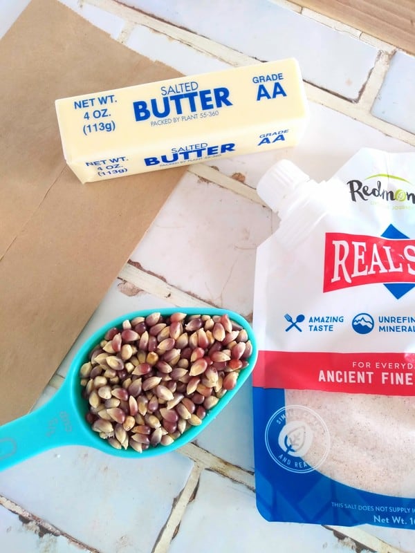 What you need for microwave popcorn includes popcorn kernels, salt, butter, and a brown paper bag sitting on a counter top.