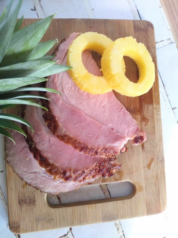 Slices of ham sitting on a wood cutting board with rings of pineapple next to it and the top of a pineapple showing.