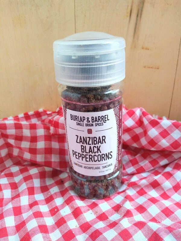 Burlap & Barrel Zanzibar Black Peppercorn grinder in front of a wood board with a red/white checkered cloth underneath.