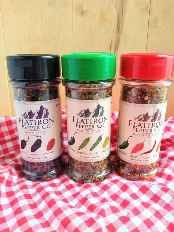 Flat Iron Pepper Company Shakers from left to right - Dark and Smoky, Hatch Valley Green, and Four Pepper Blend in front of a wood board with a red/white checkered cloth underneath.