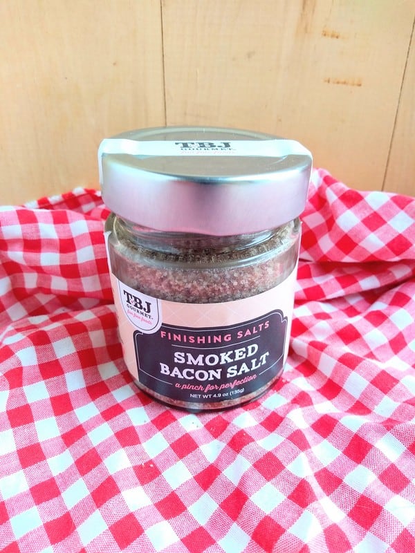 TBJ Gourmet Smoked Bacon Finishing Salt in front of a wood board with a red/white checkered cloth underneath.