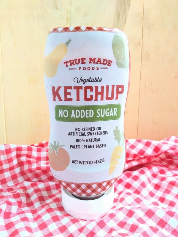 True Made Foods No Added sugar Vegetable Ketchup in front of a wood board with a red/white checkered cloth underneath.