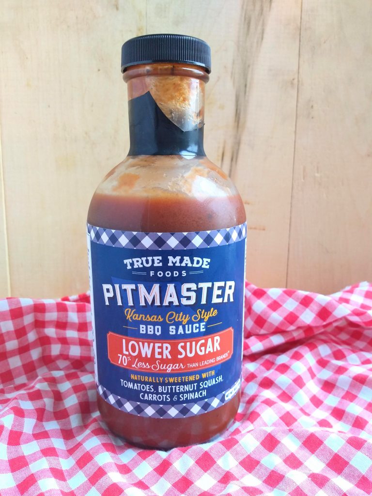 True Made Foods Lower Sugar BBQ Sauce in front of a wood board with a red/white checkered cloth underneath.