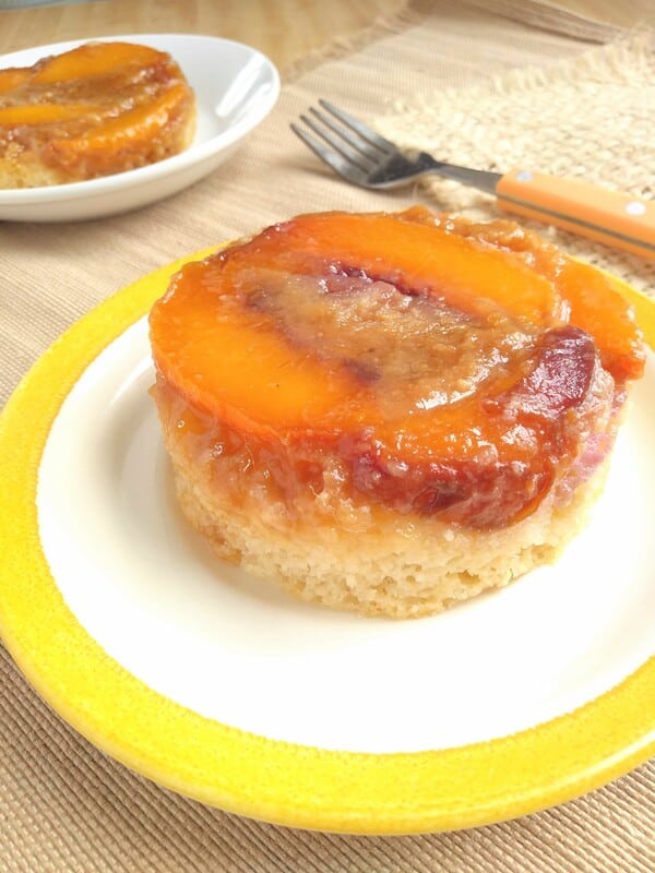 Alton Brown's Upside Down Peach Cakes sitting on a white plate with a yellow border with a fork and a second cake sitting in the background