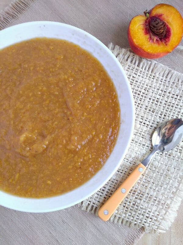 Peach applesauce in a large white bowl on top of a piece of burlap with a orange handled spoon to the right of the bowl and a peach cut in half.