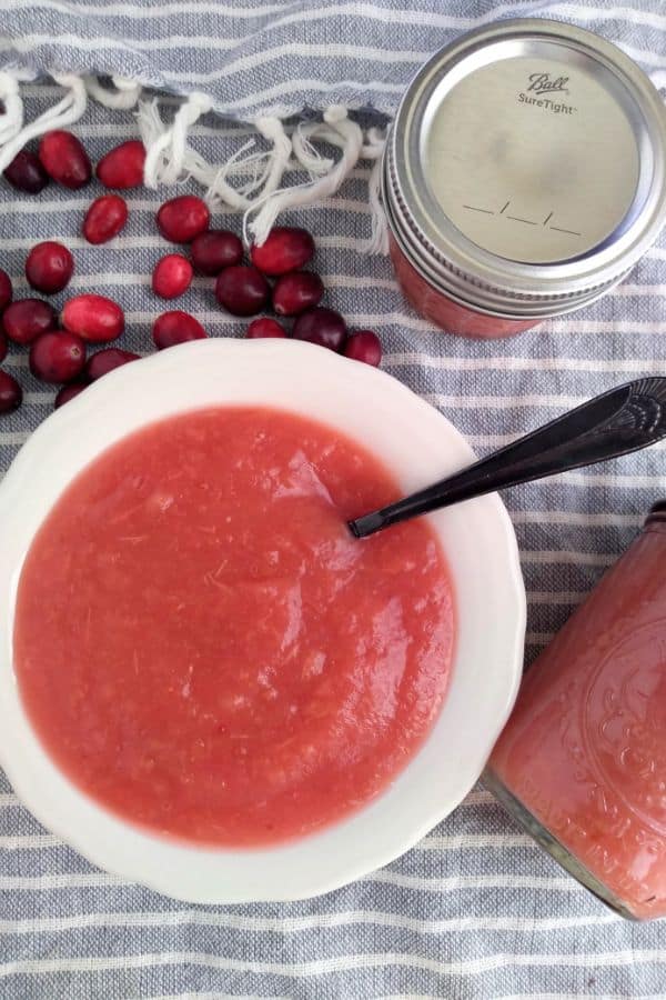 A bowl of red cranberry applesauce with a black spoon sticking out on top of a gray and white stripped towel with jars of applesauce and fresh cranberries around the edge. 