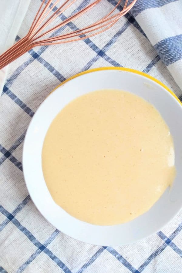 Mustard sauce in a white bowl with a yellow plate underneath. A blue striped white towel is underneath with a bronze color whisk on top of it.