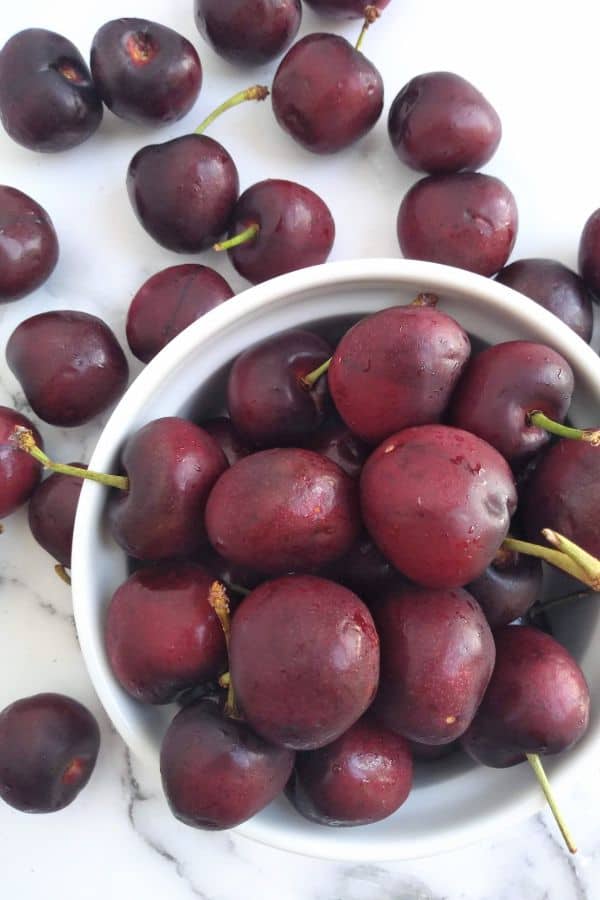 Bing cherries inside a white ramekin with cherries surrounding it on top of a white background