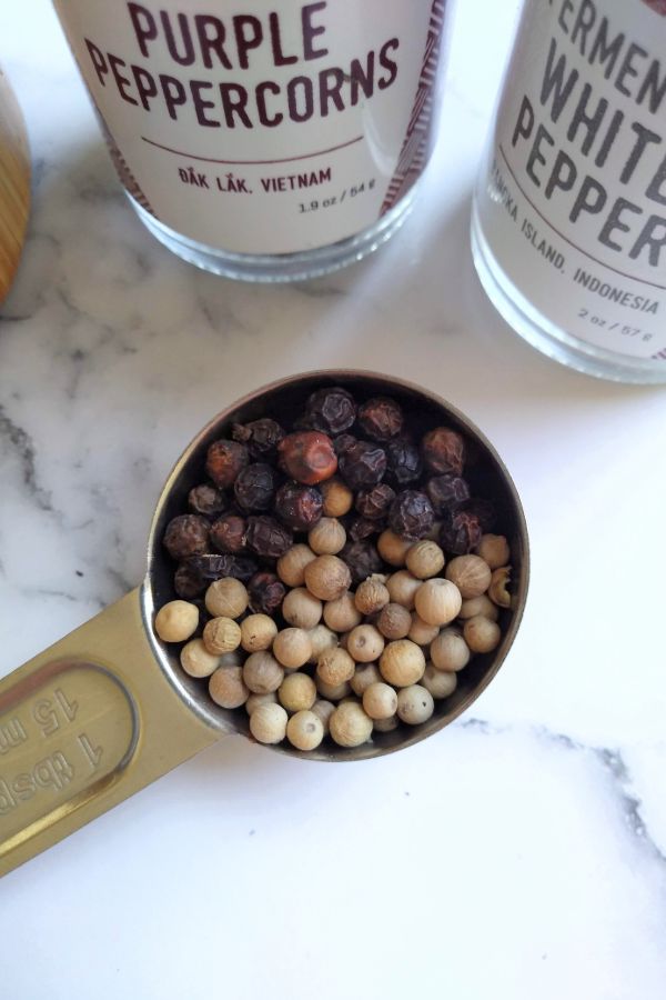 Burlap & Barrel Purple Peppercorns and Fermented White Pepper are divided in a tablespoon measuring spoon with the grinders in the background