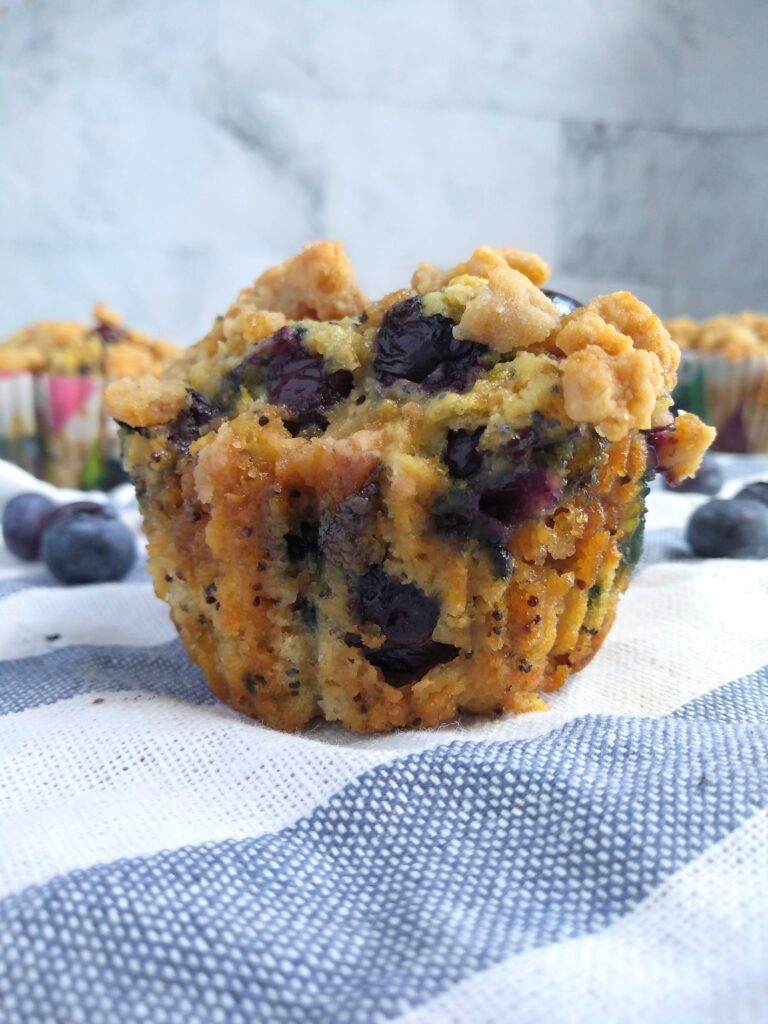 A blueberry sour cream muffins with poppy seed and lemon sitting on a blue and white striped towel with additional muffins in the background.