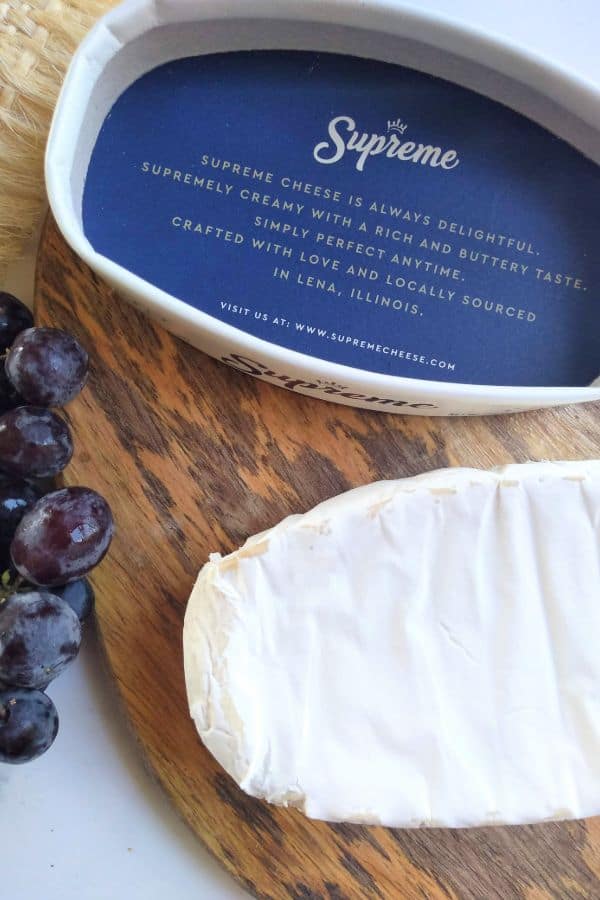 Supreme brand Brie cheese sitting on a cutting board with some grapes with the lid of the cheese upside down