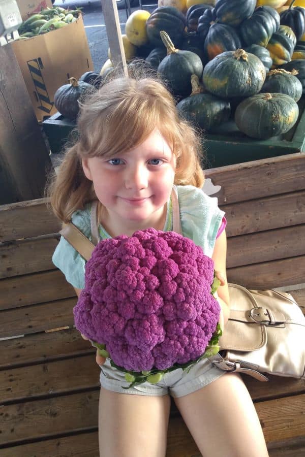 My daughter at the farmer's market holding a large head of purple cauliflower.