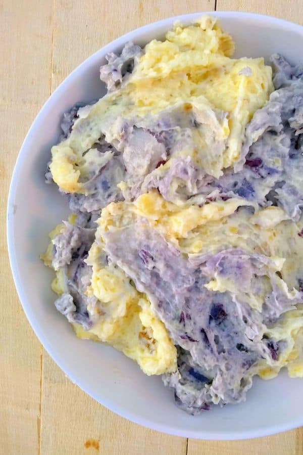 A bowl of gold mashed potatoes swirled with purple mashed potatoes sitting on a wood board.